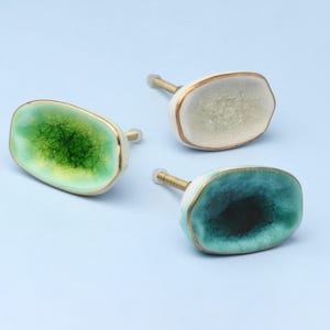 May include: Three decorative cabinet knobs in green, white, and teal. Each knob has a unique, crackled pattern and a gold-coloured trim. The knobs are mounted on a light blue background.