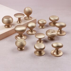 May include: A collection of twelve cabinet knobs in a matte gold finish. The knobs feature various shapes, including smooth rounded tops, flat circular tops, and ribbed cylindrical designs, each mounted on a circular base for attachment.