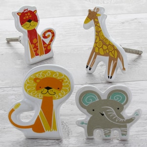 Op de afbeelding: Vier keramische ladegrepen met cartoon-dierontwerpen. De grepen zijn wit met kleurrijke dierillustraties. De dieren zijn een leeuw, een giraffe, een olifant en een tijger.