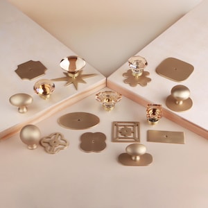 Puede incluir: Una colección de herrajes para muebles que incluye varios pomos de metal dorado y placas decorativas. El conjunto presenta pomos redondos lisos, pomos de cristal transparente facetado y placas metálicas con formas geométricas, florales y de estrella.
