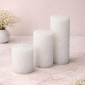May include: Three white pillar candles of varying heights. The candles have a textured surface and a single wick at the top. The candles are arranged on a light-coloured surface, with a vase of white flowers in the background.