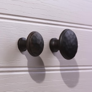 May include: Two round cabinet knobs with a dark, matte, hammered metal finish. The knobs feature a textured, dimpled surface and a central stem that attaches to a flat, light-colored, horizontally paneled wooden surface with visible grain.