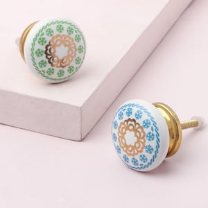 May include: Two white ceramic cabinet knobs with brass fittings. One features a green and gold floral pattern, the other a blue and gold design. The knobs are set on a light pink surface.