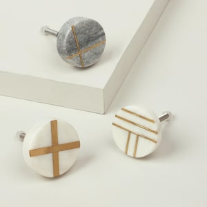 Peut inclure: Trois boutons de meuble décoratifs. L'un est en marbre gris avec un motif en croix doré. Deux sont blancs avec des motifs géométriques dorés. Chaque bouton a une vis argentée.
