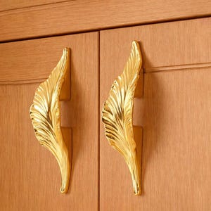 May include: Two gold-colored leaf-shaped cabinet handles mounted on a light brown wooden cabinet. The handles have a detailed, textured design, and the cabinet doors have a vertical wood grain pattern.
