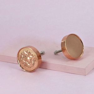 Peut inclure: Deux boutons de meuble ronds de couleur cuivre. L'un des boutons présente un motif floral en relief, tandis que l'autre a une surface lisse et unie. Chaque bouton est doté d'une vis argentée pour l'installation. Les boutons sont présentés sur une surface rose clair sur fond rose pâle.
