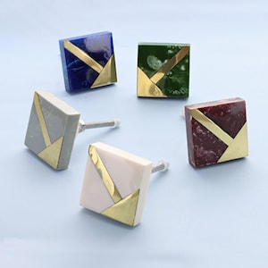 May include: Five square cabinet knobs in shades of blue, green, grey, white, and burgundy. Each knob has a gold-coloured geometric design. The knobs are made of a solid material and have a silver screw for installation.