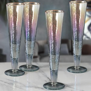 May include: Four iridescent champagne flutes with a textured surface and gold rims. The flutes have a tall, slender design and sit on a round base. The glass reflects a range of colours, including pink, blue, and yellow.