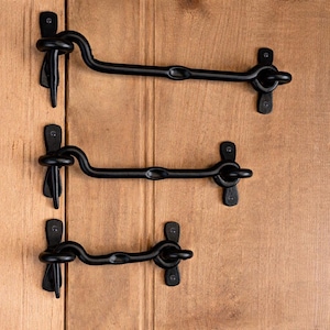 May include: Three black iron gate latches fixed to a wooden surface. Each latch has a curved hook and a matching eye for secure closure. The latches are finished in matte black, offering a rustic appearance. The wooden surface has vertical grain.