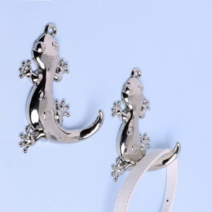 May include: Two shiny silver gecko-shaped hooks. The hooks have a loop at the top for hanging and are attached to a white strap. The geckos have detailed feet and a curved tail. The background is a light blue.