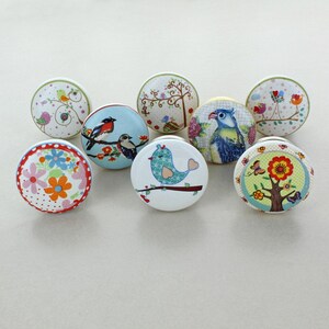 May include: A collection of eight ceramic cabinet knobs with colorful bird and floral designs. Each knob features a unique illustration, including birds on branches, flowers, and trees, set against various background colors. The knobs are round and have a metal base.