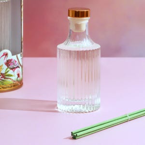 May include: A clear glass diffuser bottle with a gold cap and a white stopper. The bottle has vertical ridges and is filled with a clear liquid. Green diffuser sticks are in the foreground. The background is a gradient of pink and purple.