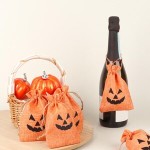 May include: Orange burlap drawstring bags with black jack-o'-lantern faces. A bottle of champagne has a bag tied around the neck. A basket with orange pumpkins is in the background. Halloween-themed decorations.