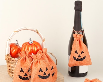 May include: Orange burlap drawstring bags with black jack-o'-lantern faces. A bottle of champagne has a bag tied around the neck. A basket with orange pumpkins is in the background. Halloween-themed decorations.