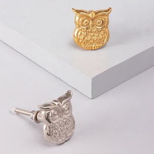 Peut inclure: Deux boutons de porte en forme de hibou, l'un doré et l'autre argenté. Le bouton doré est sur une surface blanche, tandis que le bouton argenté est sur une surface blanche avec un fond blanc.