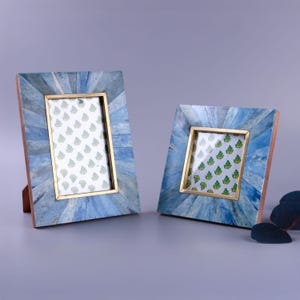 May include: Two decorative picture frames. The frames have a blue mosaic design with gold-coloured inner frames. The pictures inside the frames have a green leaf pattern on a white background. The frames are standing upright.