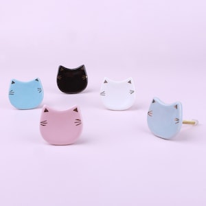 Könnte beinhalten: Fünf Keramik-Schubladengriffe in Katzenform in verschiedenen Farben: Schwarz, Weiß, Hellblau und Rosa. Die Griffe haben goldene Akzente für Augen und Schnurrhaare.