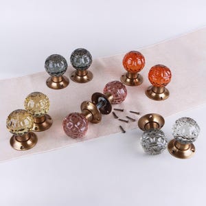 May include: A collection of round, faceted glass door knobs in various colours including clear, yellow, pink, orange, and grey. Each knob features a metallic bronze-toned circular base with screw holes and includes several small metal screws.