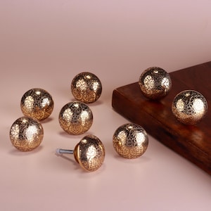 May include: Gold-coloured cabinet knobs with an ornate floral design. The knobs are spherical and have a screw attached to one of them. The knobs are arranged on a light pink surface and a wooden board.