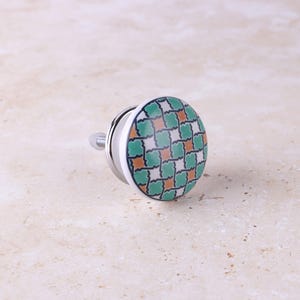 May include: A round ceramic cabinet knob featuring a geometric pattern of interlocking green, white, and orange shapes. The knob has a smooth, glossy surface and is attached to a silver-toned metal bolt and base for mounting onto furniture.