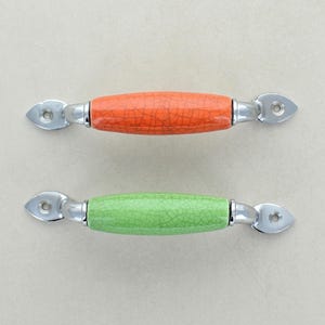 May include: Two cabinet pulls with a crackle finish. One is orange, the other is green. Both have silver-toned metal hardware. The pulls are oval-shaped and have a decorative design on the ends.