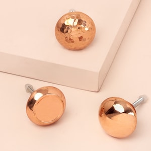 Könnte beinhalten: Drei gehämmerte Kupfer-Schrankknöpfe mit glänzender Oberfläche. Die Knöpfe sind rund und haben einen leicht erhöhten Rand.