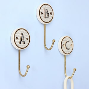 May include: Three decorative wall hooks with a white circular face, each displaying a letter: A, B, and C. The letters are black, set against a mosaic-style background. The hooks are gold-colored and mounted on a light blue wall.