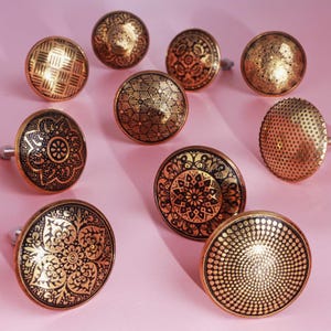 Könnte beinhalten: Eine Sammlung dekorativer Schrankknöpfe mit aufwendigen Designs. Die Knöpfe sind hauptsächlich goldfarben mit schwarzen Details und weisen Muster wie geometrische Formen, Blumenmotive und Punktmuster auf. Die Knöpfe sind rund und haben einen Metallschaft zur Befestigung.