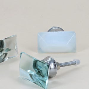 Op de afbeelding: Drie decoratieve kastknoppen met facetgeslepen, rechthoekige glazen vlakken. Eén knop is lichtblauw en de andere twee zijn helder. Elke knop heeft een zilverkleurige metalen basis en schroef.