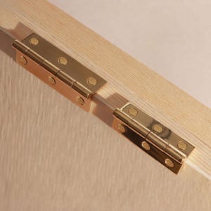May include: Two polished brass butt hinges are mounted onto a light-colored wooden surface. Each hinge features a central barrel and four flat-head screws with slotted drives, securing the metal plates firmly into the grain of the wood.