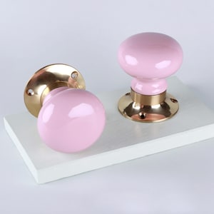 May include: Two pink ceramic door knobs with brass bases. The knobs have a rounded shape and a glossy finish. The brass bases are circular with screw holes. The knobs are displayed on a white surface.