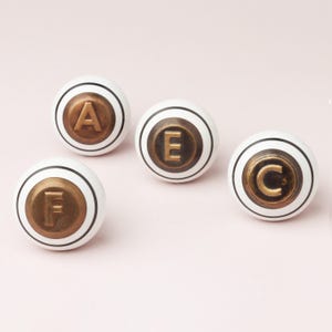 Puede incluir: Cuatro pomos de armario blancos con detalles metálicos de color bronce. Cada pomo presenta una letra diferente: A, E, F y C. Los pomos tienen una franja negra y están sobre un fondo rosa claro.
