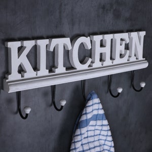 Può includere: Un cartello da cucina in legno bianco con la parola "KITCHEN" in lettere in rilievo. Il cartello ha quattro ganci in metallo nero con punte in ceramica bianca.