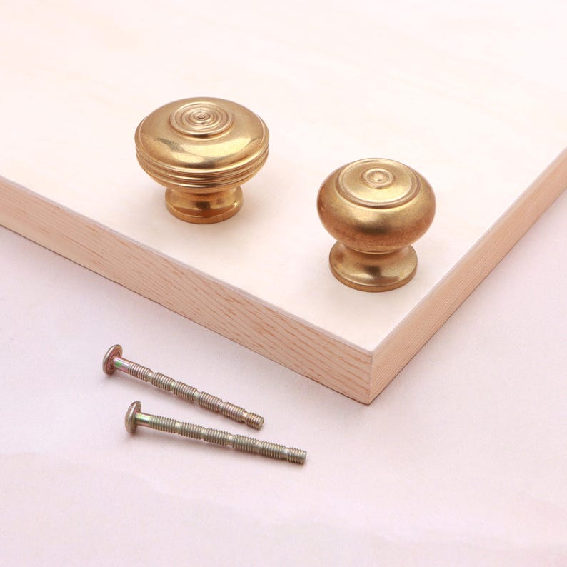 May include: Two round brass cabinet knobs with a concentric circle design on the top surface and a tapered base. The knobs feature a polished metallic finish and are accompanied by two matching threaded metal bolts for installation purposes.