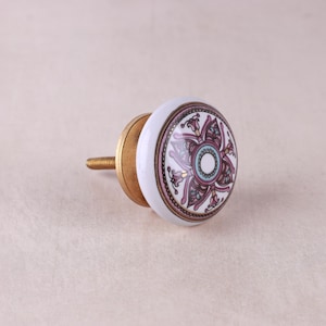Könnte beinhalten: Ein runder Keramik-Möbelknopf mit weißem Untergrund und einem dekorativen Blumenmuster in Rosa, Türkis und Dunkelbraun. Der Knopf verfügt über einen messingfarbenen Metallsockel und eine Gewindeschraube, die an der Rückseite zur Montage herausragt.