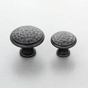 Può includere: Due pomelli per mobili in stile antico con una texture martellata. I pomelli sono grigio scuro con una parte superiore arrotondata e una base a piedistallo. Uno è più grande dell'altro.