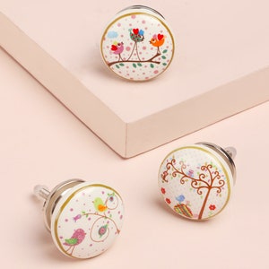 Può includere: Tre pomelli per armadi in ceramica bianca con bordo dorato. Ogni pomello presenta un diverso design di uccelli e alberi su uno sfondo bianco con pois rosa.