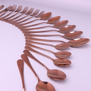 Puede incluir: Un juego de cubiertos de oro rosa, que incluye cucharas y tenedores, dispuestos en una línea curva sobre un fondo blanco.