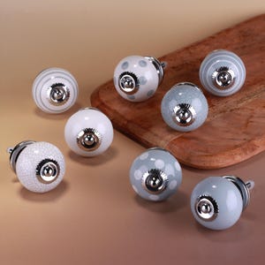 Puede incluir: Una colección de pomos decorativos para armarios en varios diseños. Los pomos presentan colores blanco, gris y azul con patrones como rayas, lunares y craquelado. Cada pomo tiene una base de metal plateado.