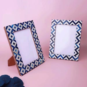 Puede incluir: Dos marcos de fotos decorativos con un patrón geométrico azul y blanco sobre un marco de madera. Un marco está en ángulo con un soporte de madera, el otro está en posición vertical. El fondo es de color rosa liso.
