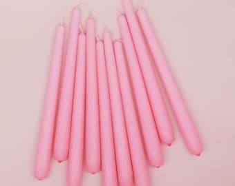 May include: A collection of eleven slender, solid light pink taper candles arranged in a fan-like formation. Each candle features a smooth, cylindrical wax body with a rounded base and a pointed top, all sharing a uniform pastel pink colour.