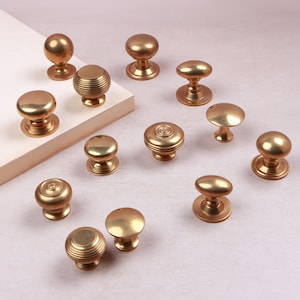 May include: A collection of twelve brass cabinet knobs in various shapes and finishes. Some knobs feature a smooth, polished surface, while others have a ribbed or concentric circular design. Each knob includes a circular base plate for mounting.