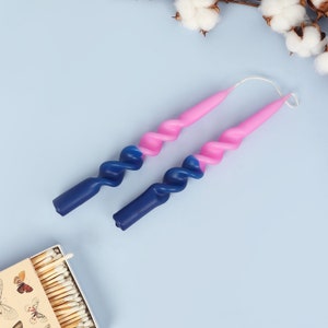 Könnte beinhalten: Zwei lange, gedrehte Kerzen mit einem rosa-blauen Ombre-Design. Die Kerzen befinden sich auf einem hellblauen Hintergrund.
