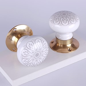 Könnte beinhalten: Zwei weiße Türknöpfe aus Keramik mit grauem Blumenmuster. Die Knöpfe haben einen Messingfuß und sind vor einem weißen Hintergrund platziert. Die Knöpfe sind rund und haben eine glatte, glänzende Oberfläche.