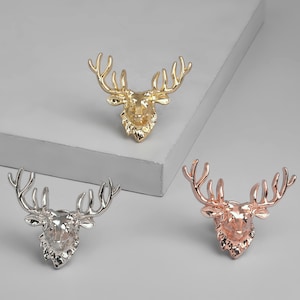 Può includere: Tre sculture decorative a testa di cervo in oro, argento e oro rosa. Ogni scultura presenta corna dettagliate e un volto strutturato. Le teste di cervo sono esposte su una superficie grigio chiaro.