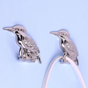 以下が含まれることがあります： 2つの銀色の鳥の形をしたフック。 羽とくちばしの詳細が施されたフックは、水色の背景に配置されています。 1つのフックが白いストラップを保持しています。 これらの装飾的なフックは、アイテムを吊るすのに適しています。
