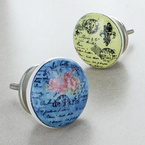 Könnte beinhalten: Zwei dekorative Schrankknöpfe mit Vintage-Design. Ein Knopf hat einen blauen Hintergrund mit rosa Blumen und Schrift, der andere einen gelben Hintergrund mit schwarzen Stempeln und Schrift. Beide haben silberne Hardware.