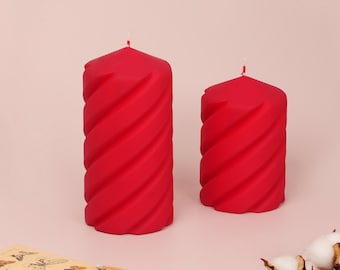 May include: Two red spiral-design candles with white wicks. The candles are different sizes and stand on a light pink surface. A box of matches with butterfly illustrations is in the lower left corner.