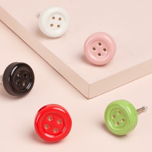 May include: Five ceramic drawer knobs shaped like buttons in various colours: white, pink, black, red, and green. Each knob has four holes and a silver screw.