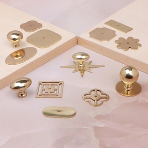 May include: A collection of polished gold-tone metal cabinet knobs and decorative backplates in various shapes including circles, squares, ovals, stars, and clover-like designs. The hardware pieces are arranged on light-coloured wooden surfaces.